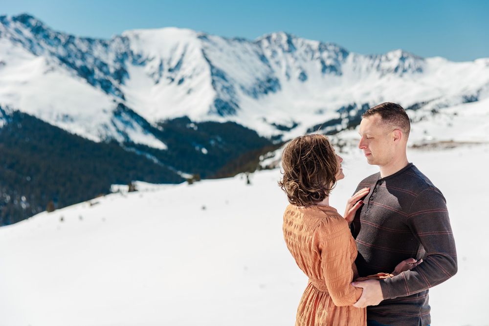 A couple shares a romantic moment on a snowy mountain overlook with snow-capped peaks in the background.