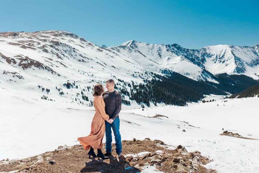 A couple stands together on a snowy mountain peak overlooking a vast winter landscape with snow-capped mountains.