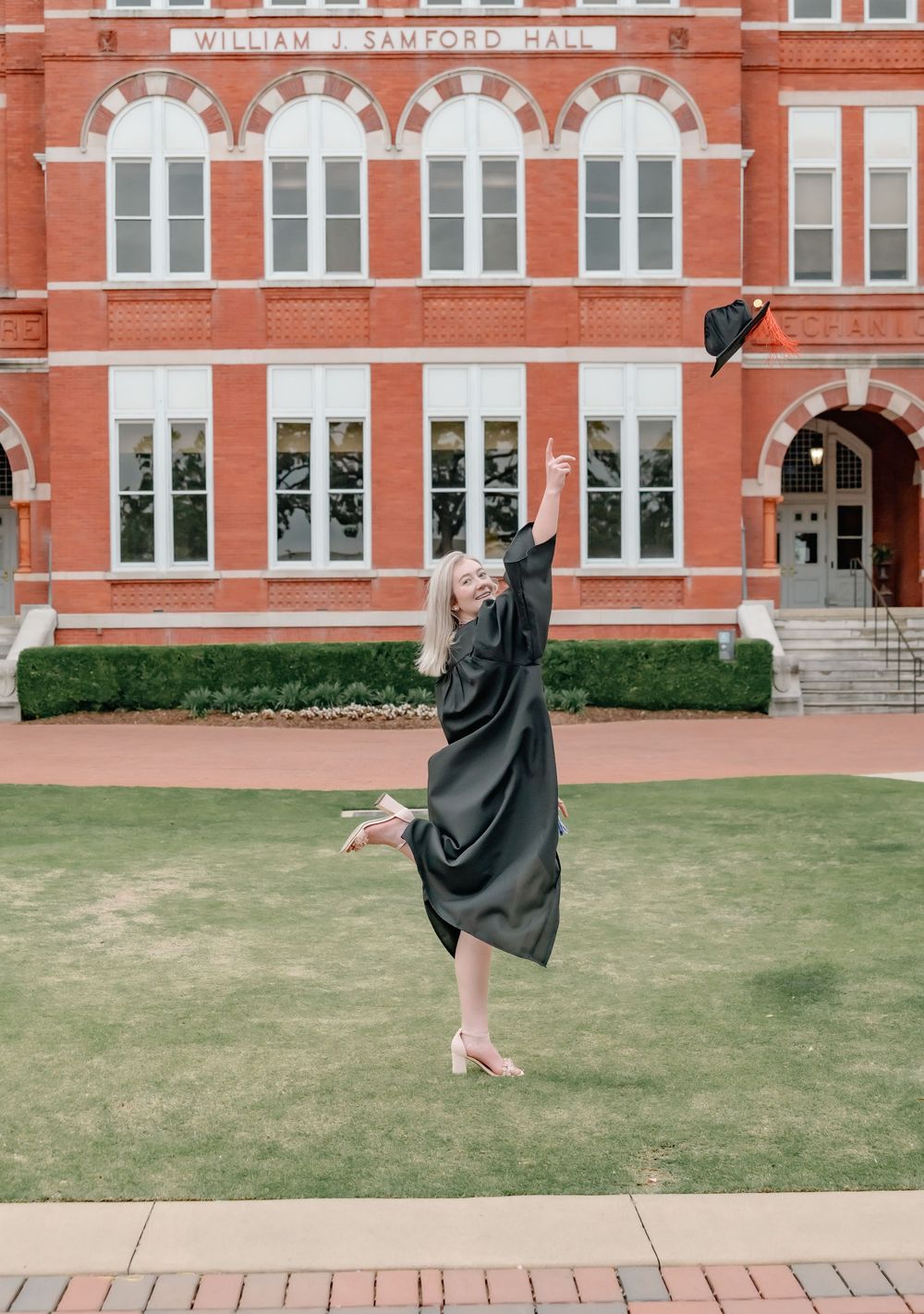 Graduate in black cap and gown jumps joyfully in front of Samford Hall at Auburn University.