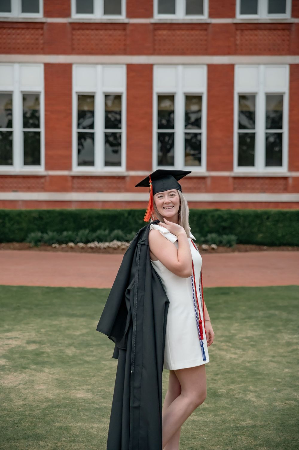 Graduate in cap and gown poses in white dress against red brick building.