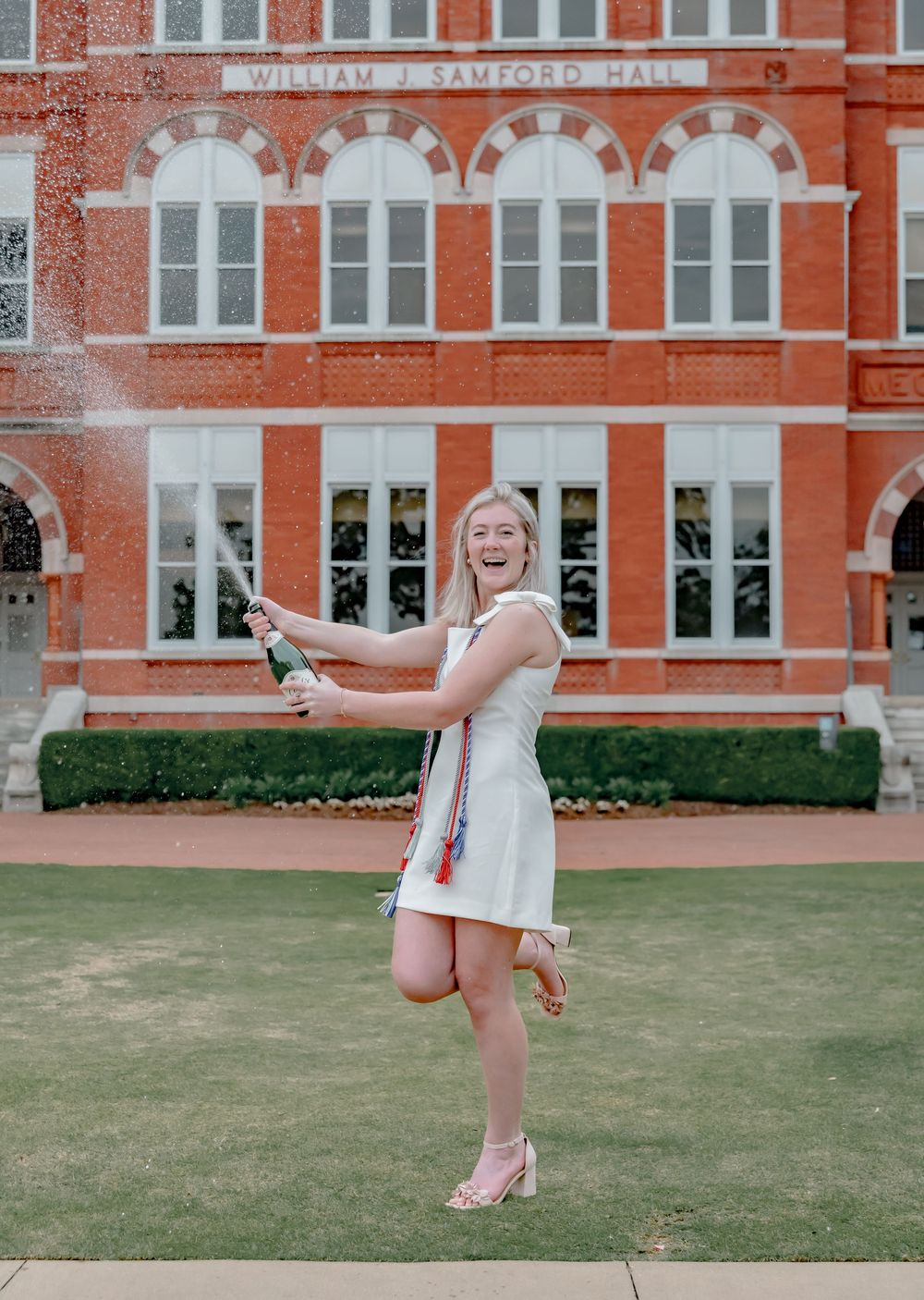 Graduate sprays champagne while celebrating in front of historic university building.