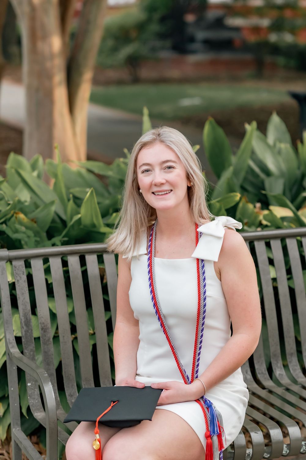 Graduate sits on bench with diploma holder wearing graduation cords and white dress.