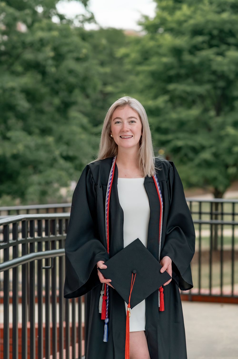 Graduate in black cap and gown poses outdoors on campus bridge during graduation photoshoot.