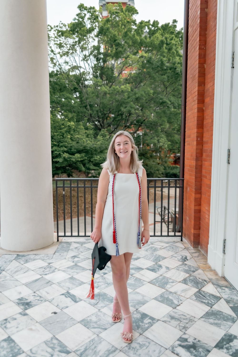 Graduate posing in a white dress on a marble balcony with columns and greenery in the background.