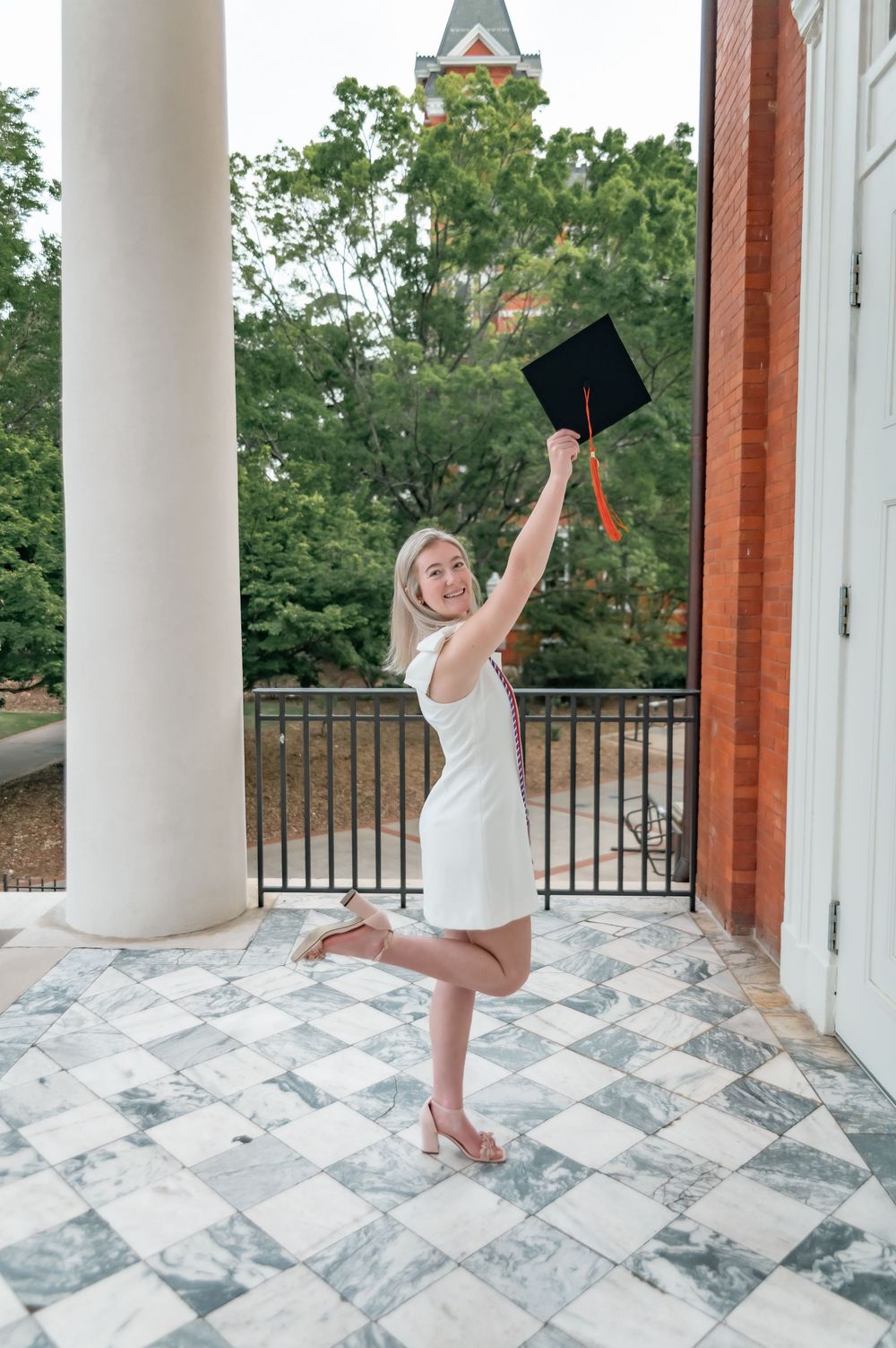 Graduate celebrating with raised graduation cap on ornate marble balcony.