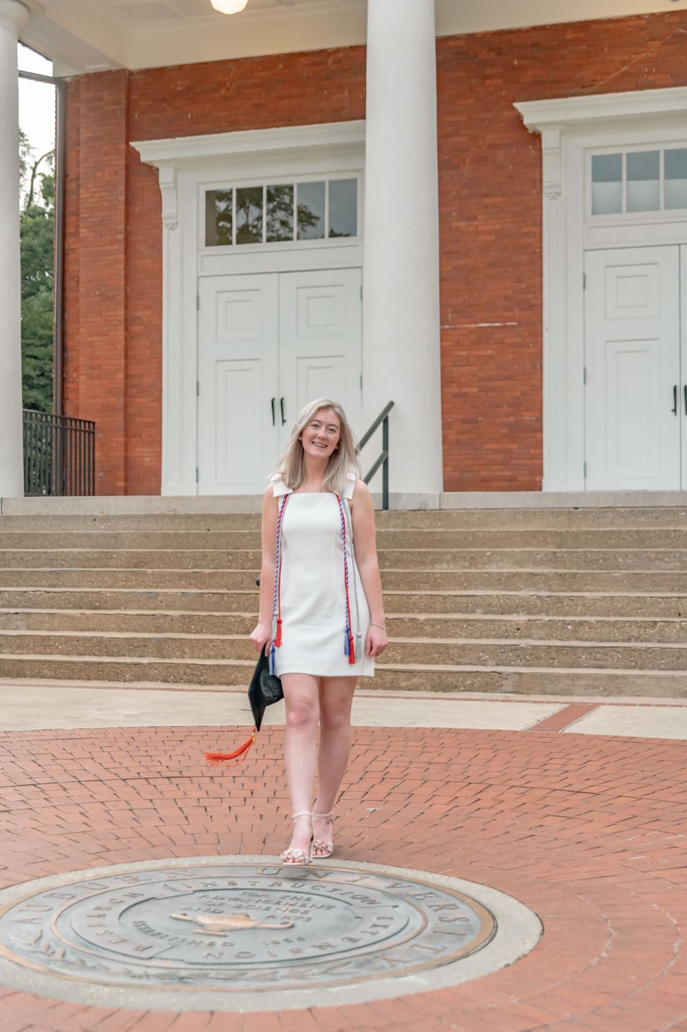 Graduate in white dress standing by brick building entrance with decorative seal on ground.