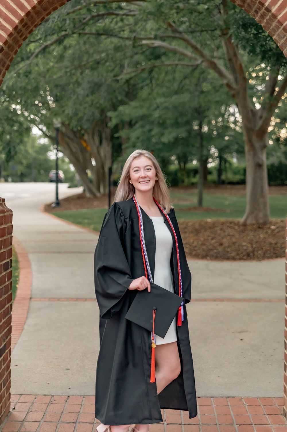 Graduate in black cap and gown poses on brick pathway with trees in background.