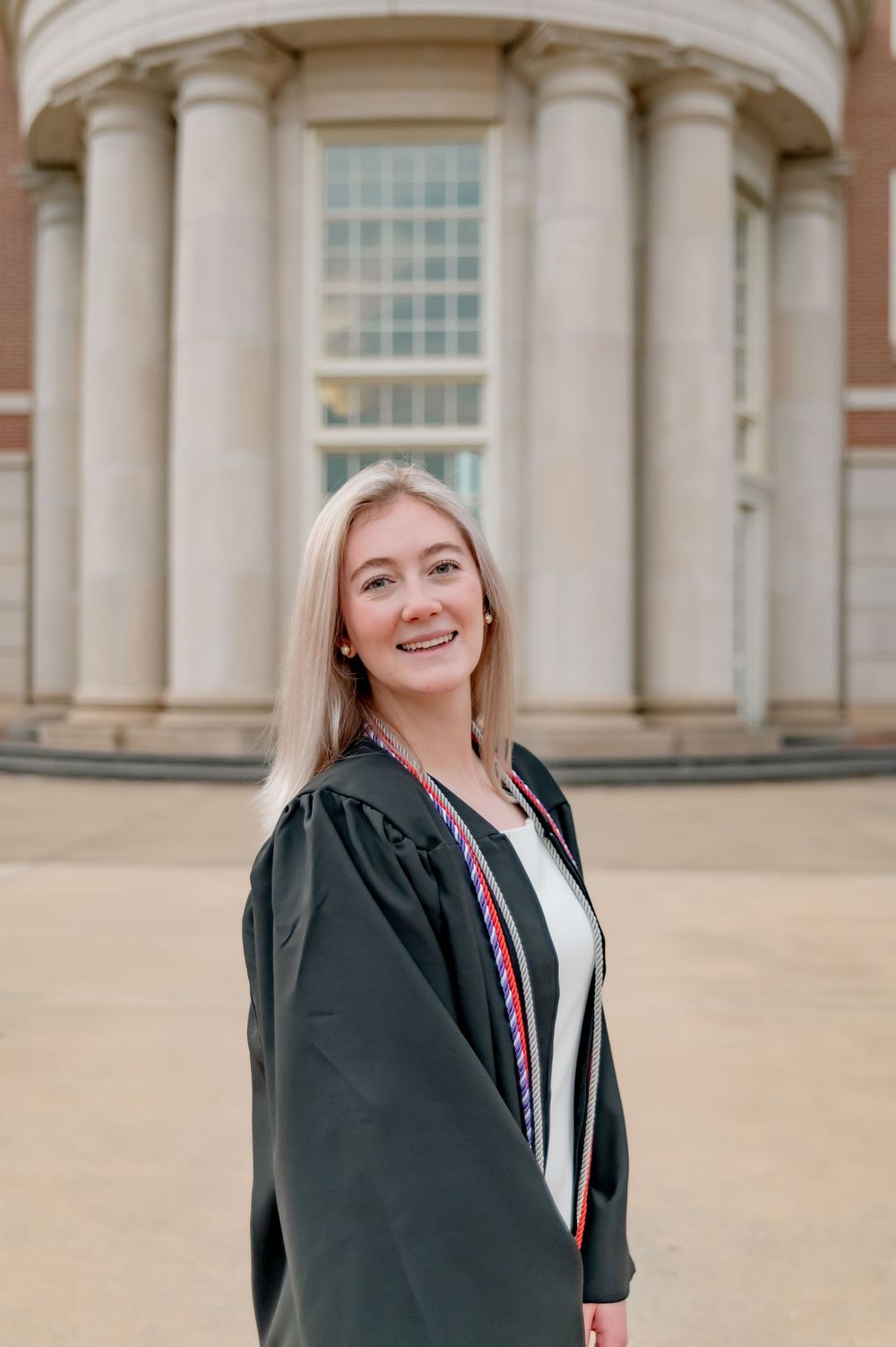 Graduate in black graduation gown poses in front of columned building entrance.
