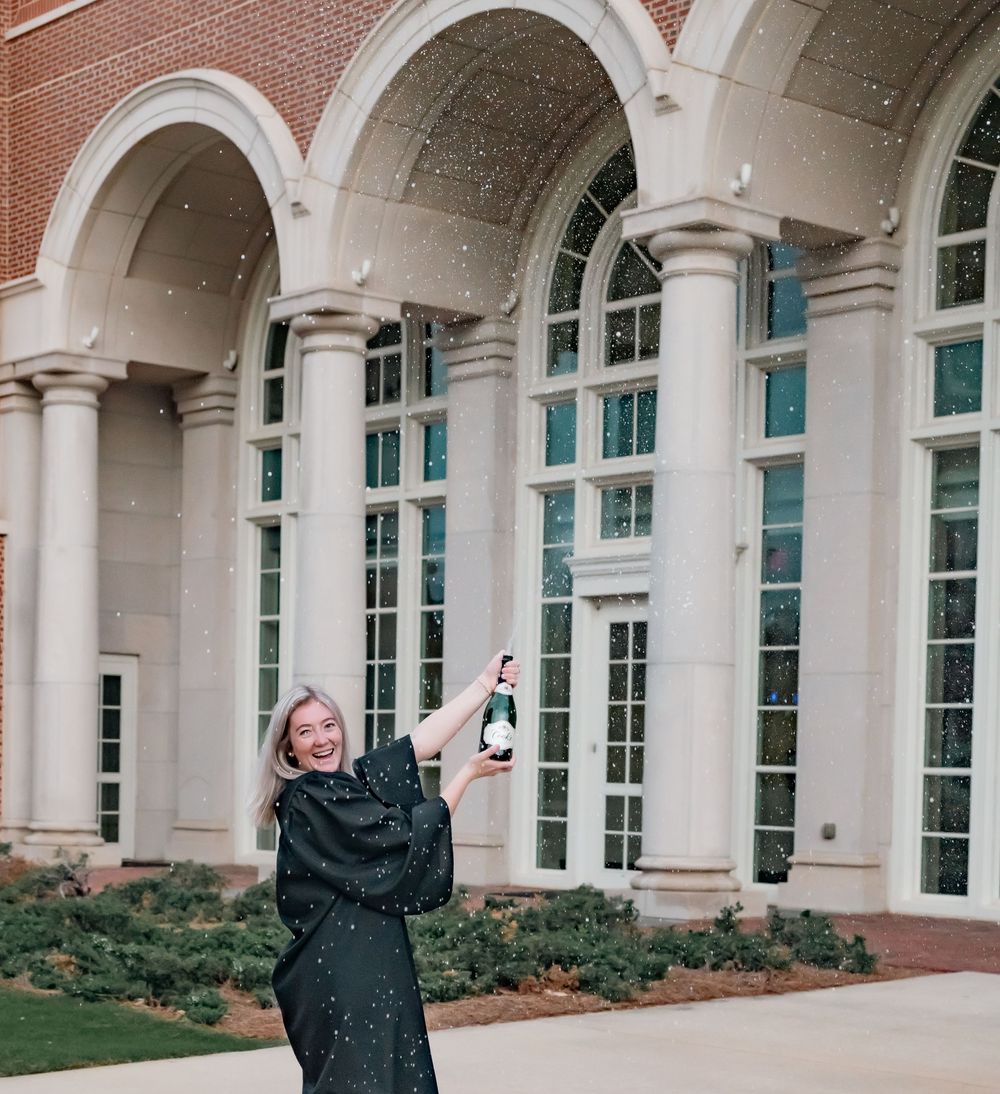 Graduate celebrating with champagne bottle in front of arched university building.