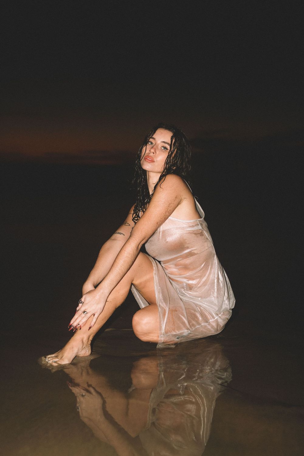 A person in a sheer dress sits on wet sand at night, creating a reflection in shallow water.