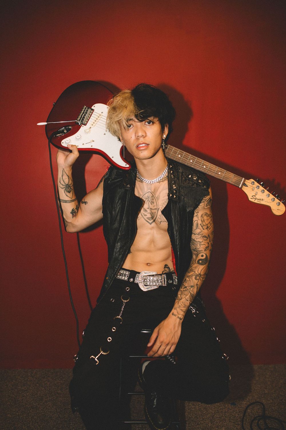 Musician in leather vest holds electric guitar against red backdrop showing tattoos and edgy style.