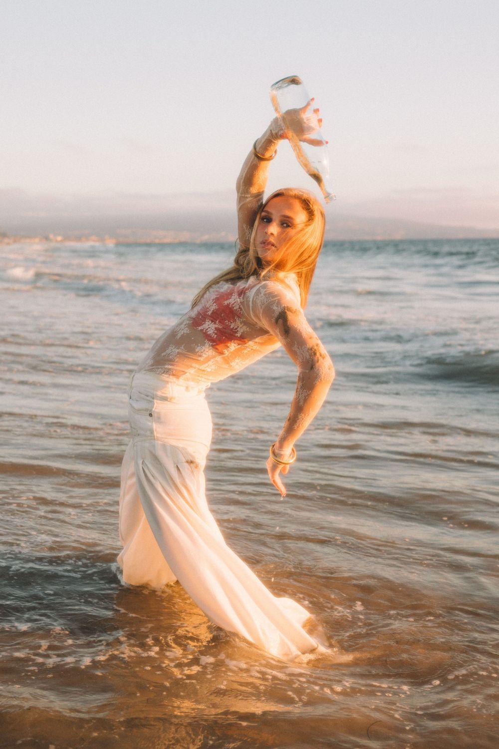 Dancer in white dress poses gracefully in ocean waves during golden hour.
