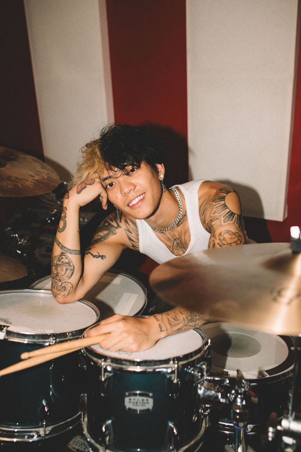 Drummer with tattoos smiles while posing with drumset in recording studio.