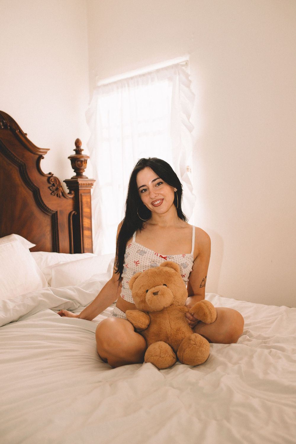 Person in white nightwear poses with teddy bear on white bedding in sunlit bedroom.
