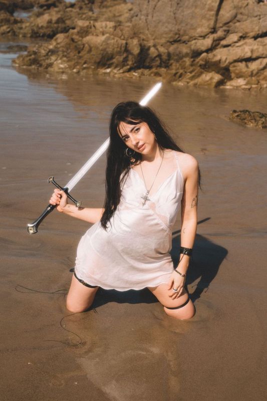 Person in white dress poses with lightsaber prop on beach rocks at sunset.