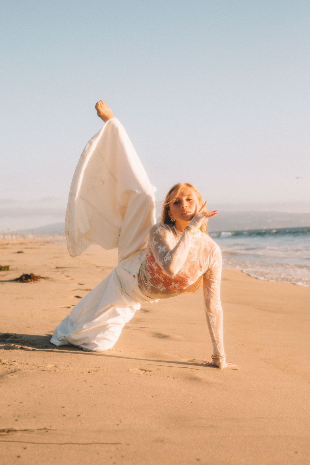 Dancing figure in flowing white dress performs graceful movement on sunny beach.