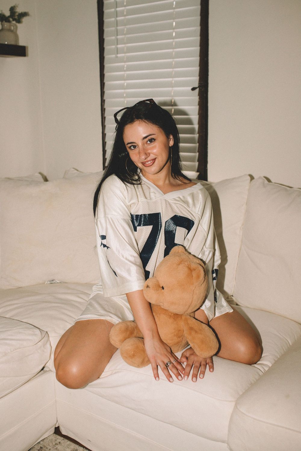 Person in white jersey lounges with teddy bear on white couch.