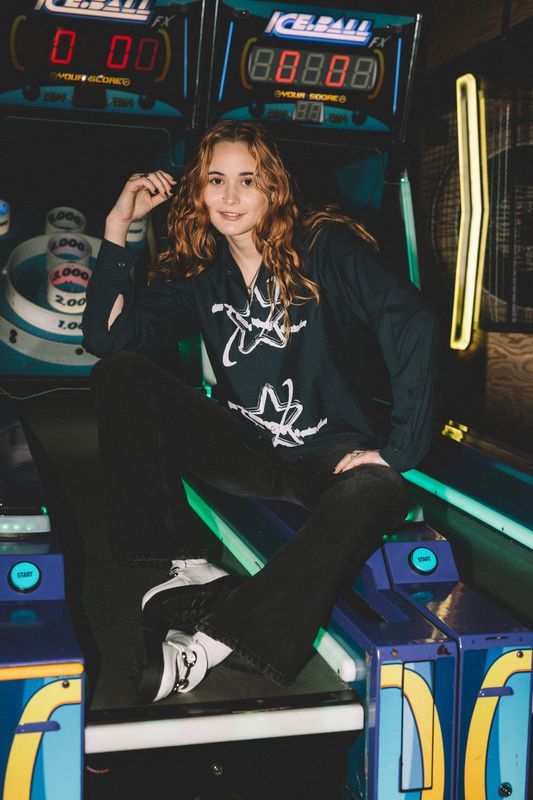 Person in dark clothing poses playfully at an arcade with illuminated game machines in the background.