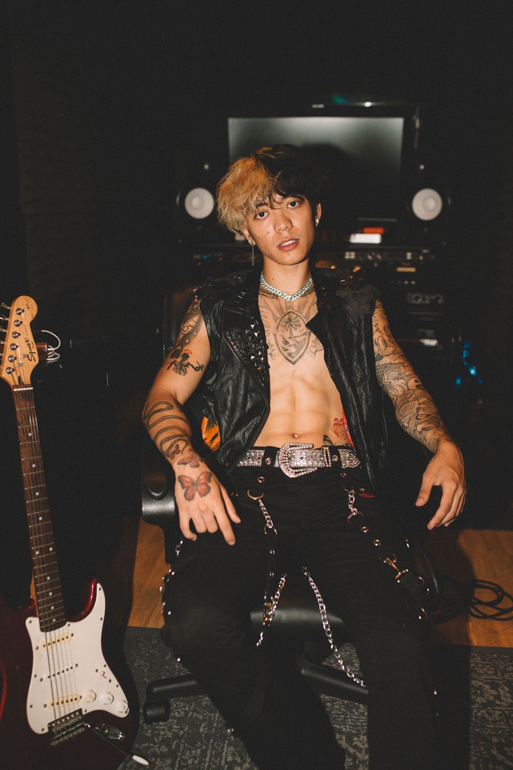 Tattooed musician in leather vest poses with electric guitar in dimly lit studio setting.