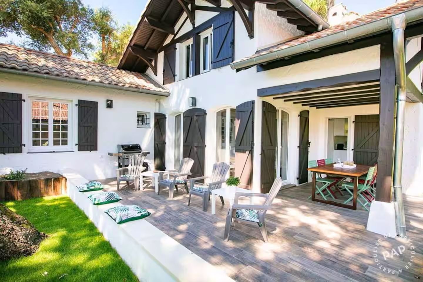 Charming Basque-style villa with white exterior and black shutters features a sunny patio with lounge chairs and garden.