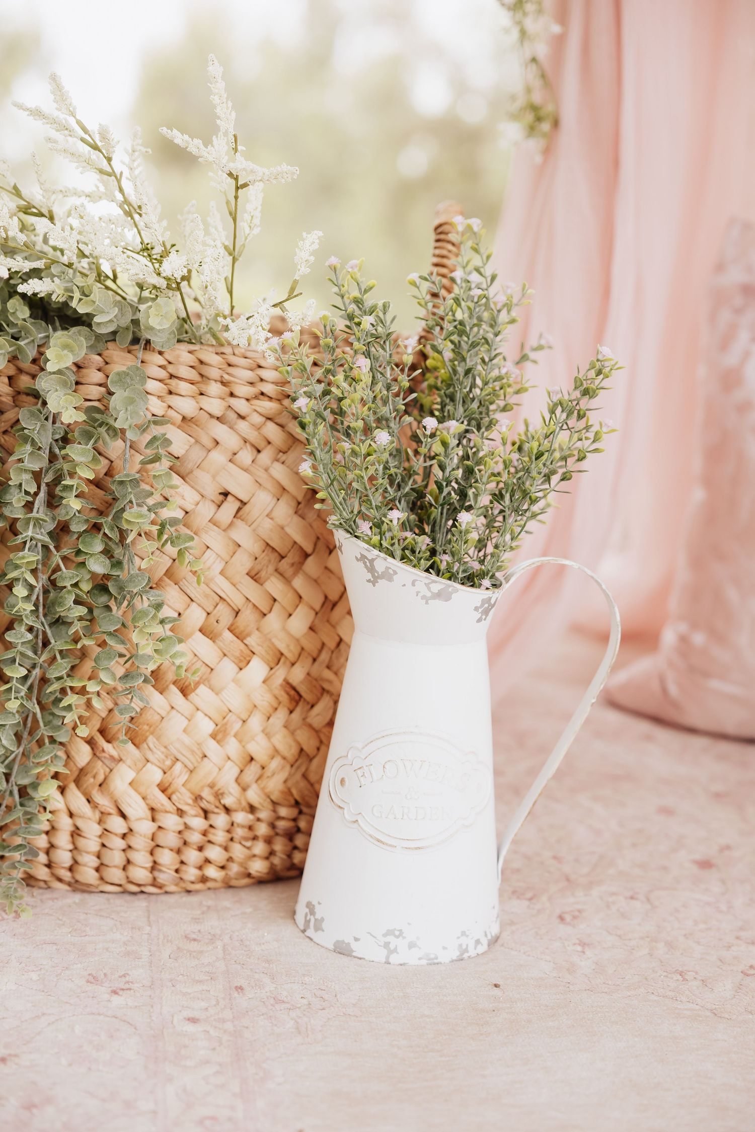 Vintage white metal pitcher filled with fresh eucalyptus sprigs next to woven basket against pink drapes.