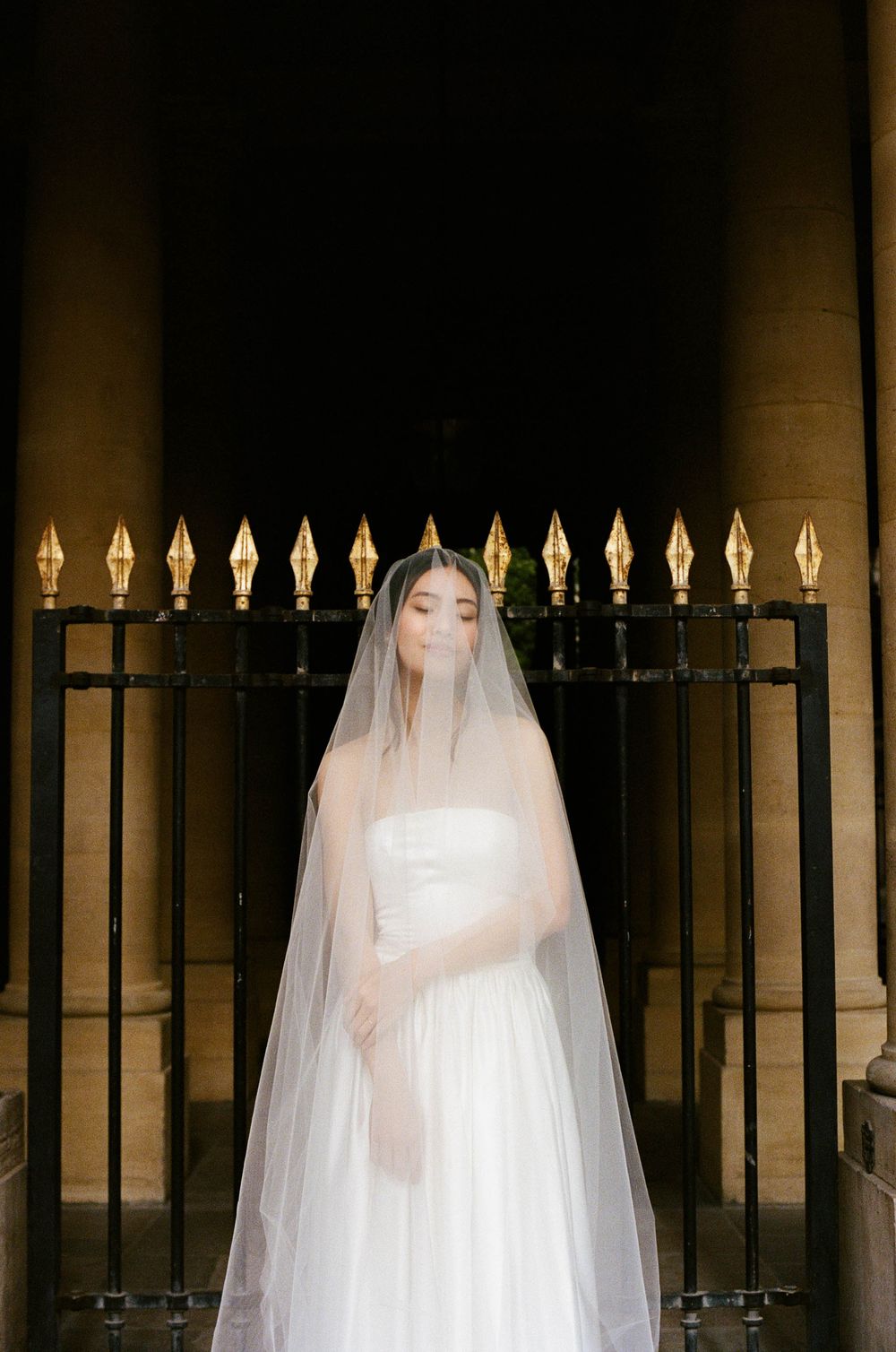 A bride in an elegant white wedding dress and flowing veil poses in front of ornate iron gates with decorative finials.