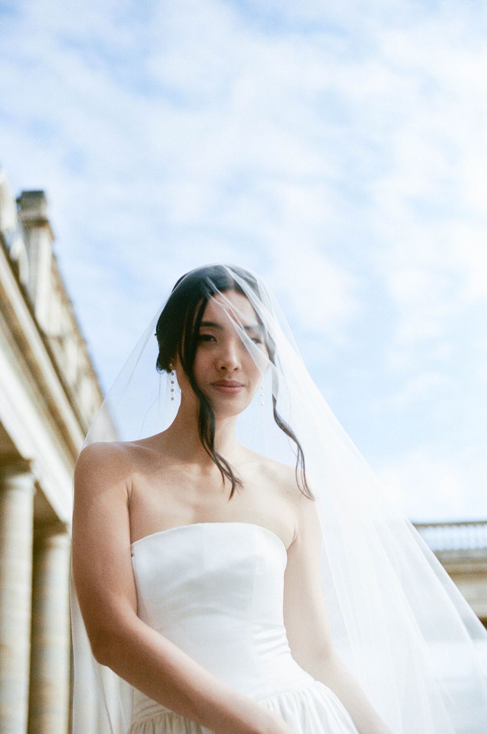 A bride in a classic white strapless wedding dress and flowing veil stands against a stone building on a sunny day.