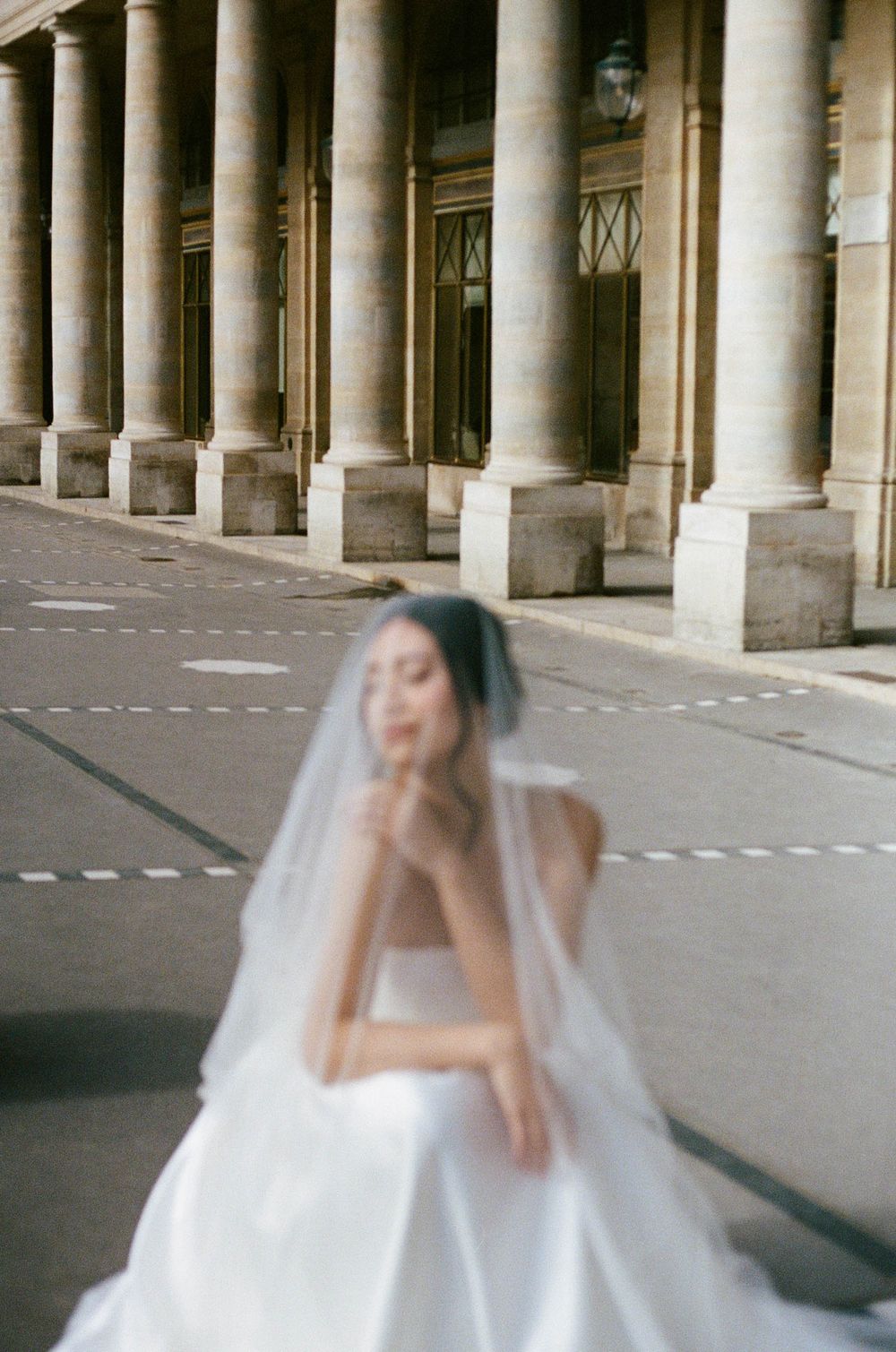 A bride in a flowing white wedding dress and veil poses in front of classical stone columns at an elegant venue.