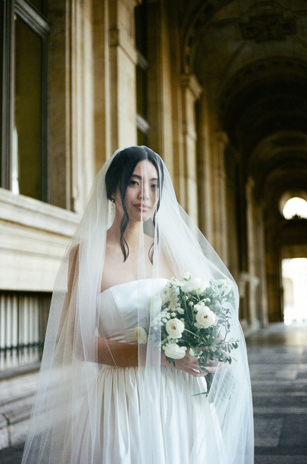 A bride in a white wedding dress and veil holds a delicate bouquet of white flowers in an elegant columned hallway.