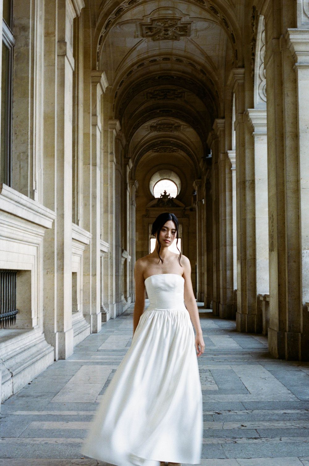 A white wedding dress is showcased in a grand archway corridor with classical architecture and stone columns.