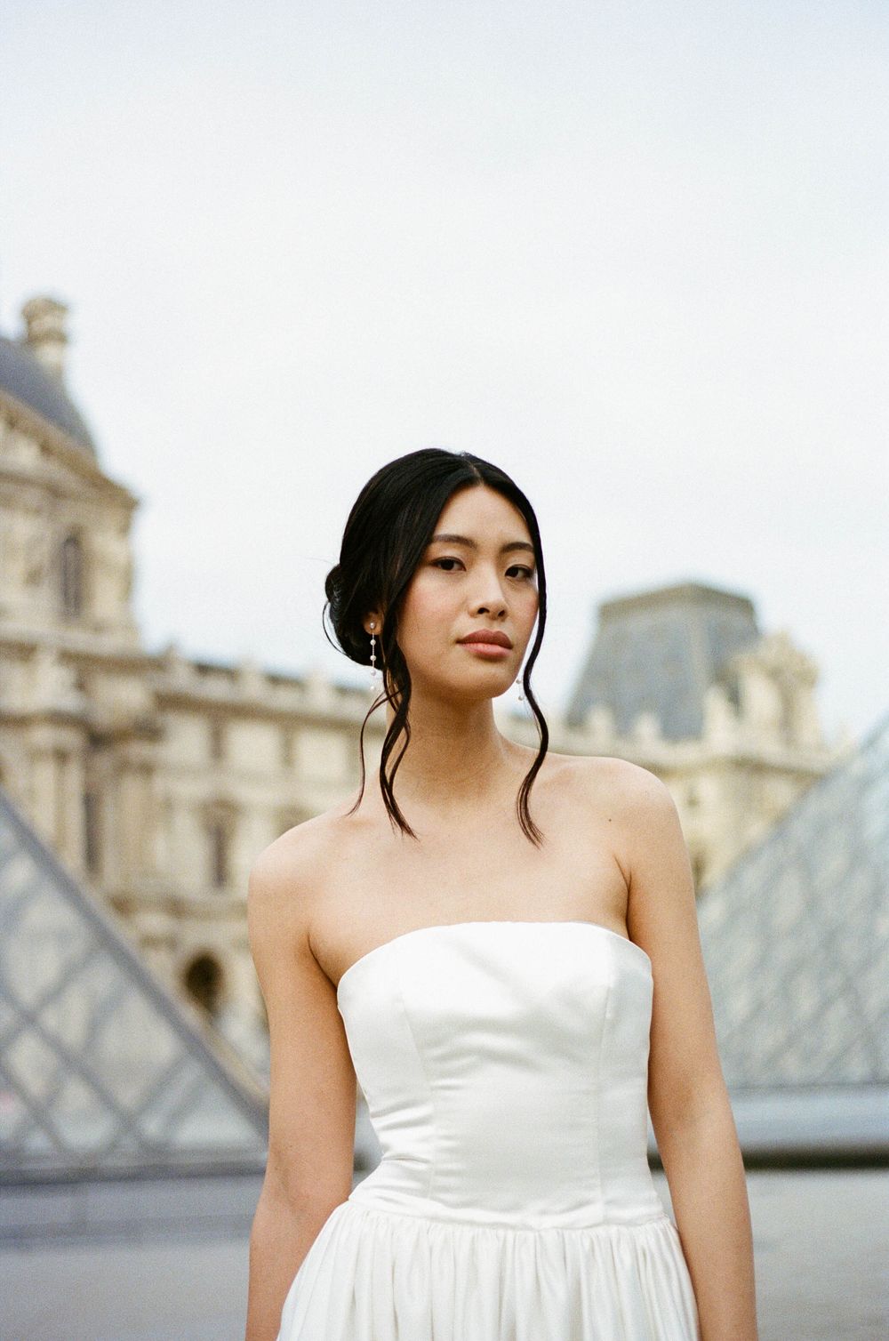Elegant strapless white dress photographed in front of the Louvre Museum and glass pyramid in Paris.