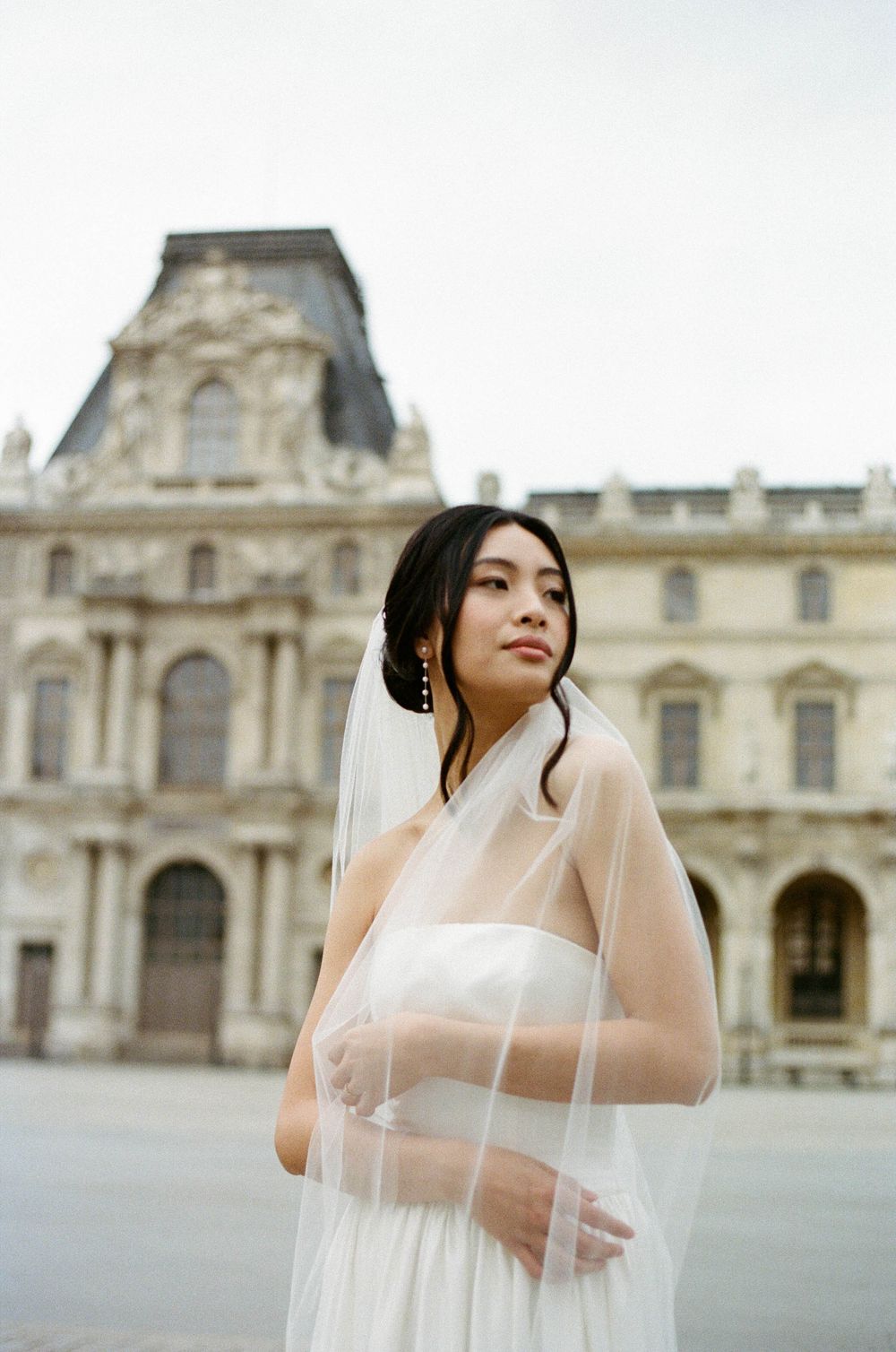 A bride in a flowing white veil poses elegantly outside the historic Louvre Museum in Paris.