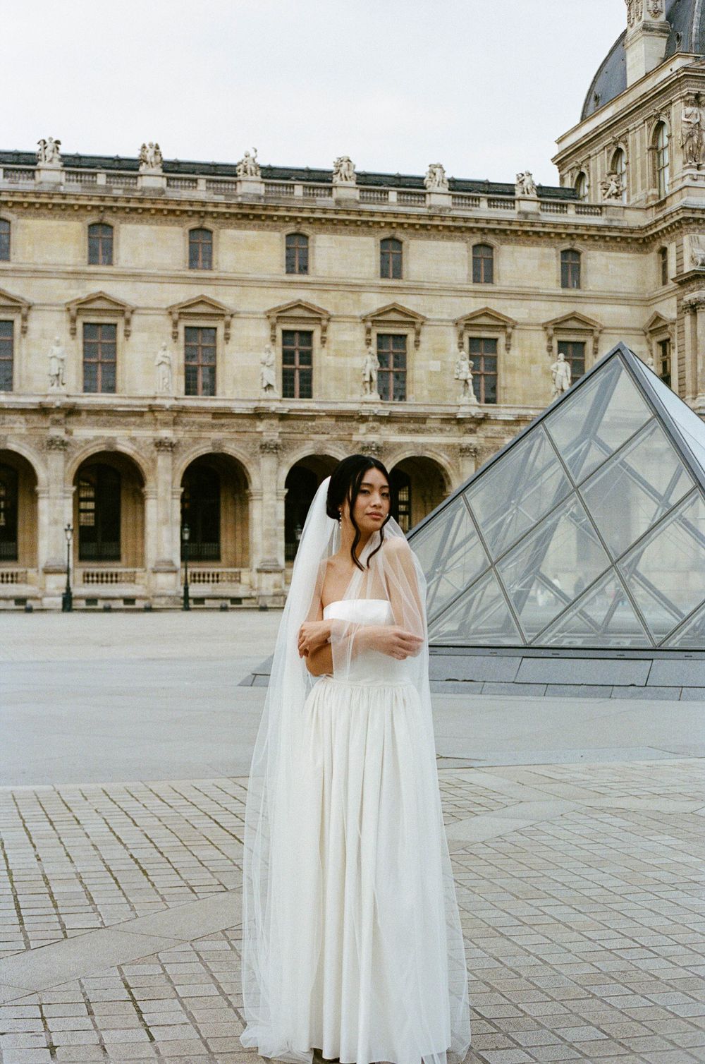 Person in elegant white wedding dress poses in front of iconic glass pyramid at the Louvre Museum in Paris, France.