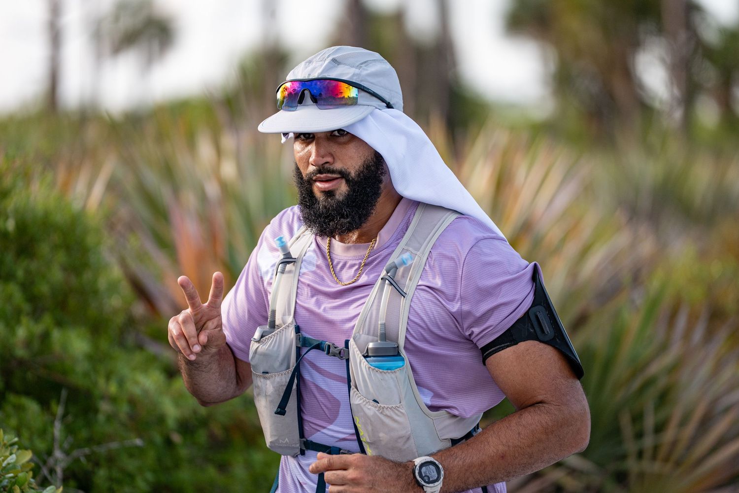 Runner wearing purple shirt, white sun protection and sunglasses gives peace sign while training outdoors among palm trees.