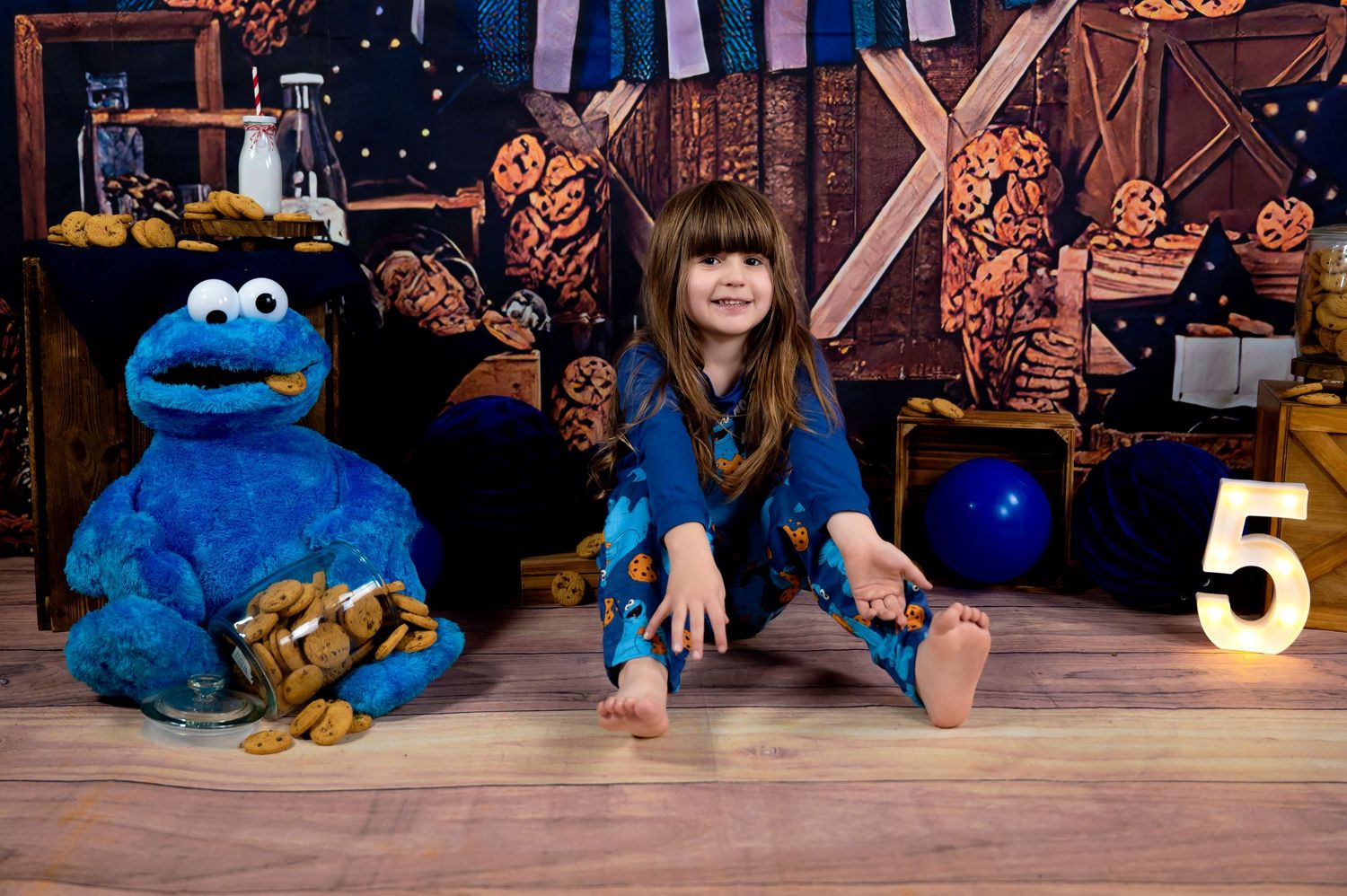 A child sits on the floor next to Cookie Monster from Sesame Street in a decorated studio backdrop setting.
