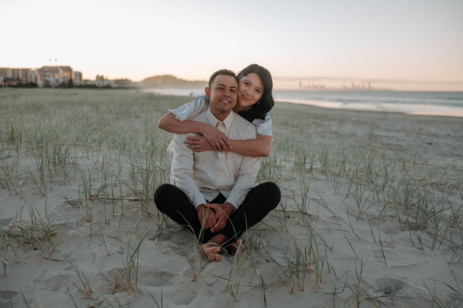 Two people share a warm embrace while sitting together on sandy dunes at sunset by the beach.