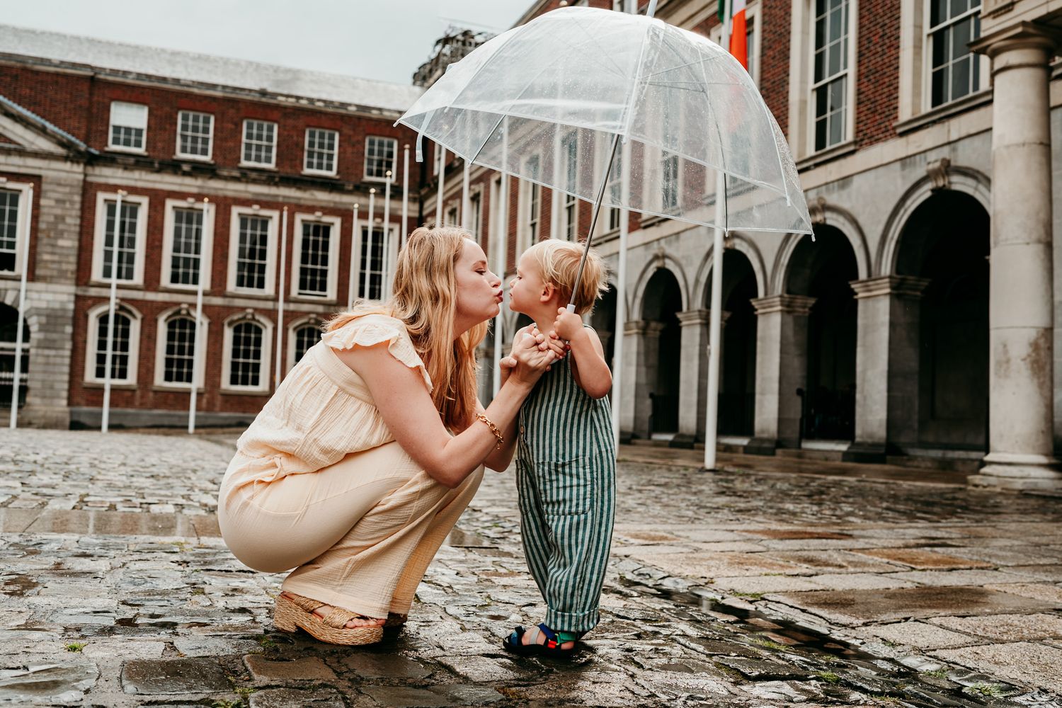 A woman and child share a tender moment under a clear umbrella in front of a historic brick building with arches.