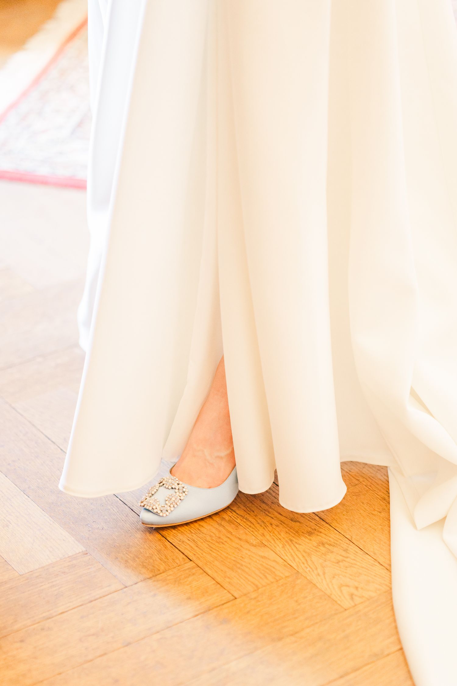 Nahaufnahme des Saums eines fließenden weißen Brautkleids und der Brautschuhe auf Holzboden.