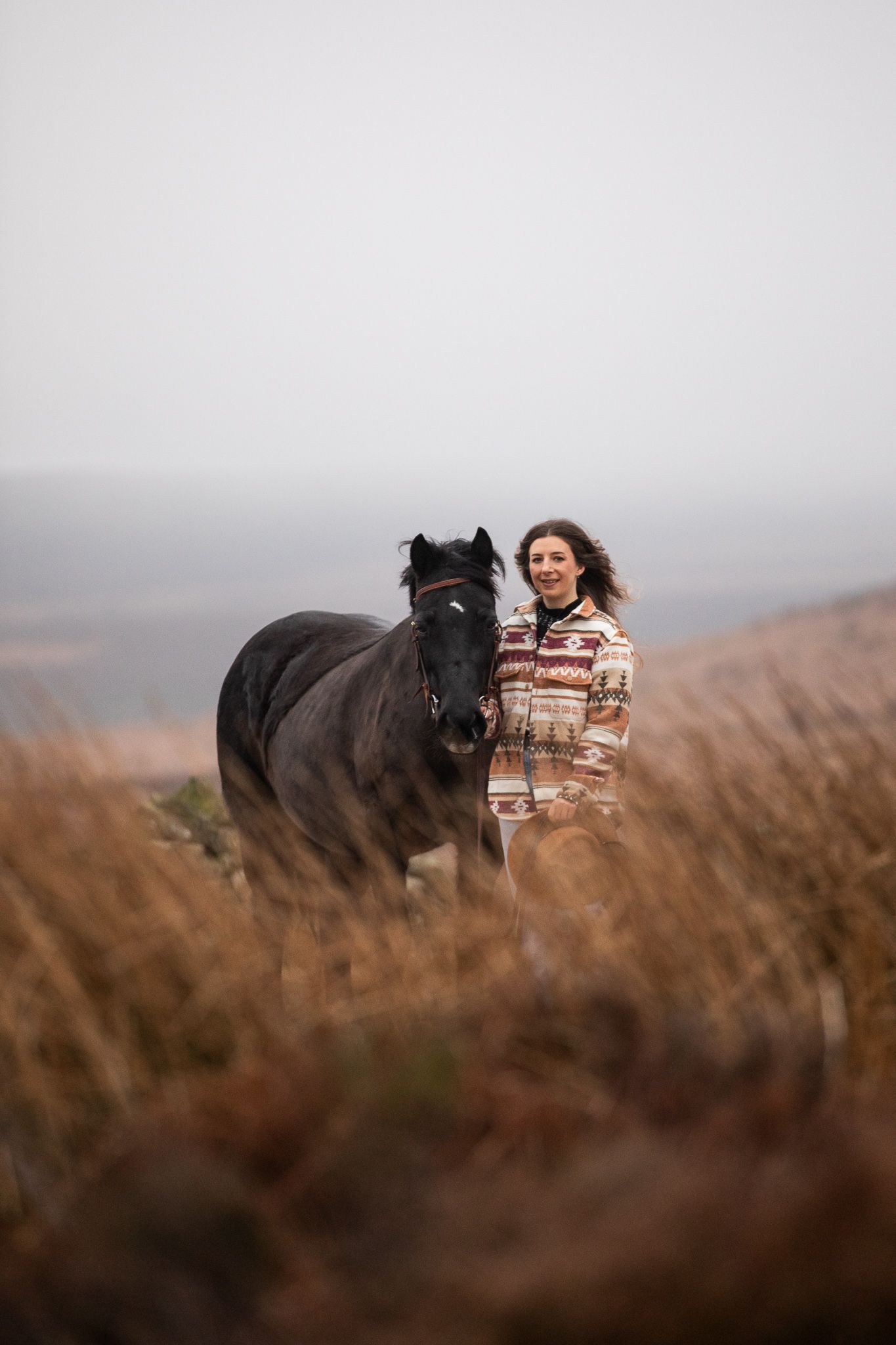 A woman in a long brown dress stands beside a majestic white horse on a rustic hillside in warm evening light.