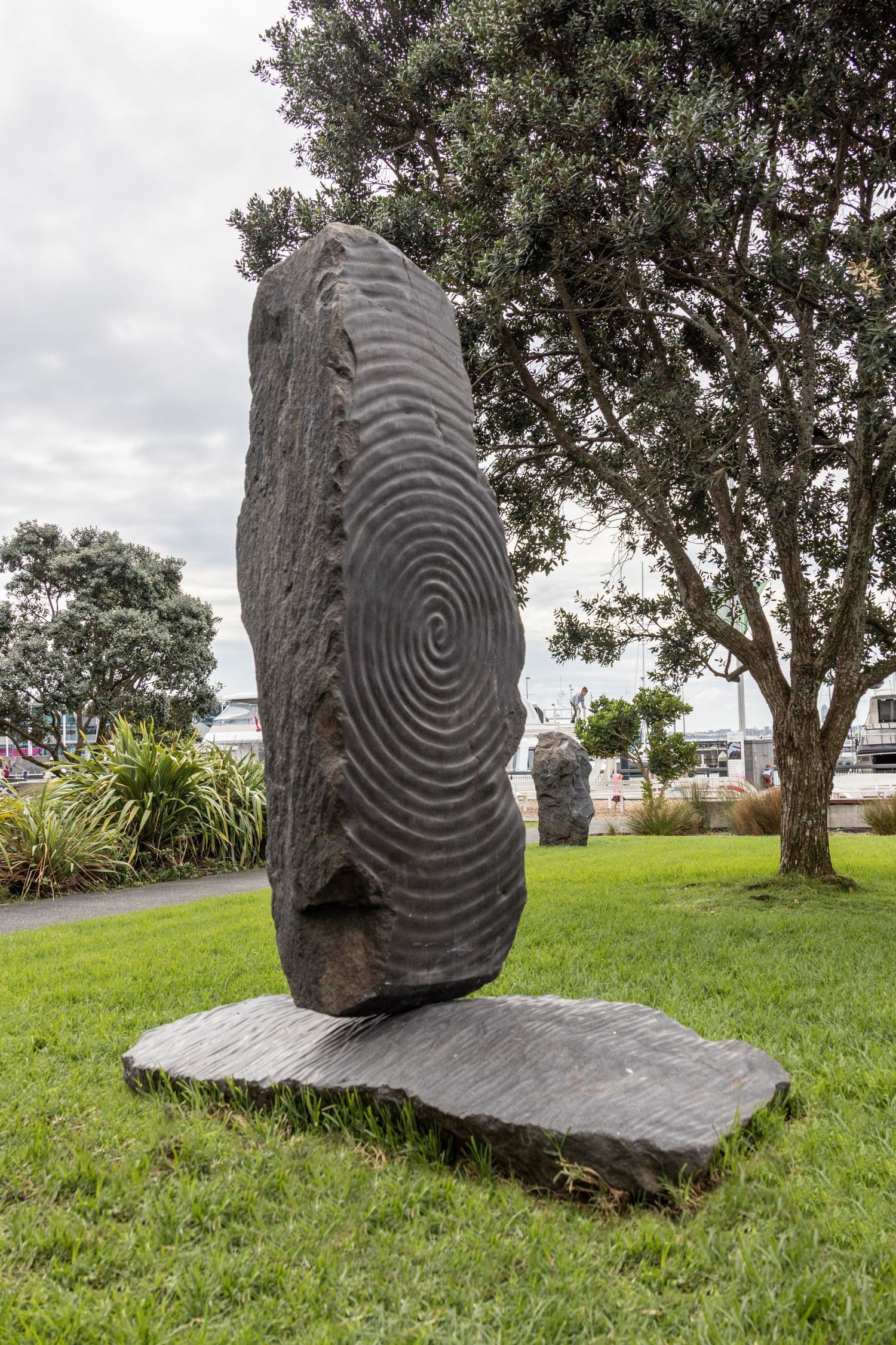 Tall textured stone sculpture standing upright on a flat base in a grassy park, with trees and cloudy sky in the background.
