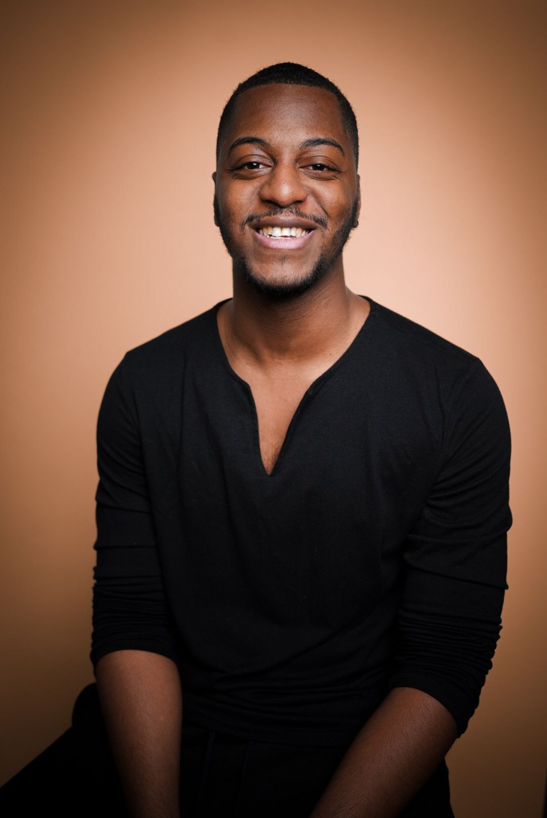 Professional portrait of someone smiling warmly while wearing a black v-neck sweater against a brown backdrop.