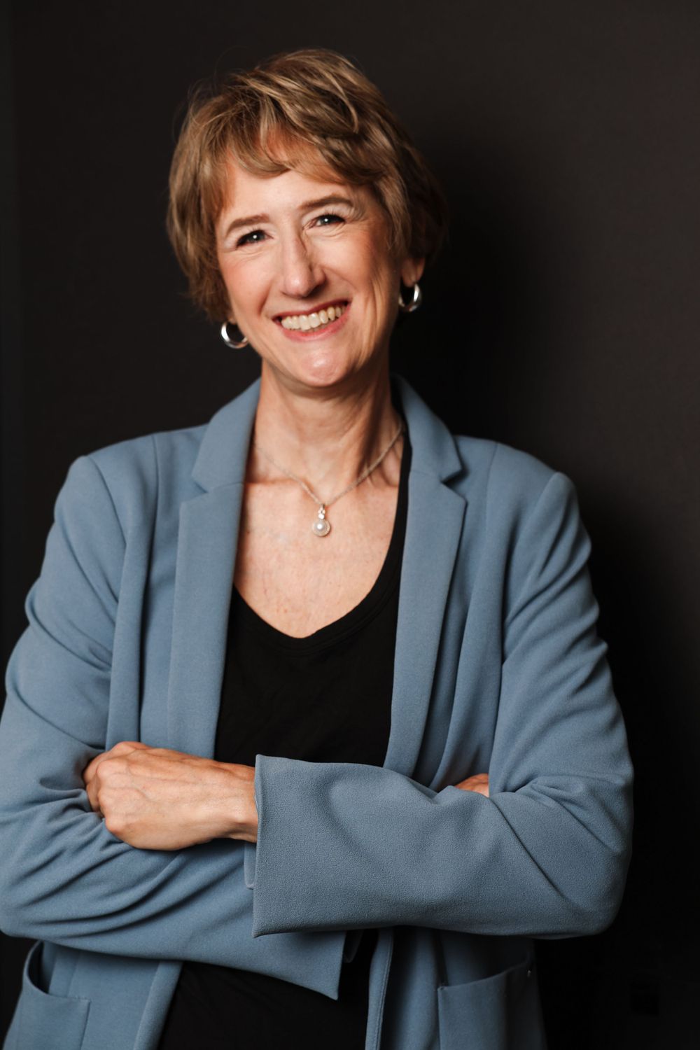 Professional portrait of a person in blue blazer and black top with short hair against dark background.