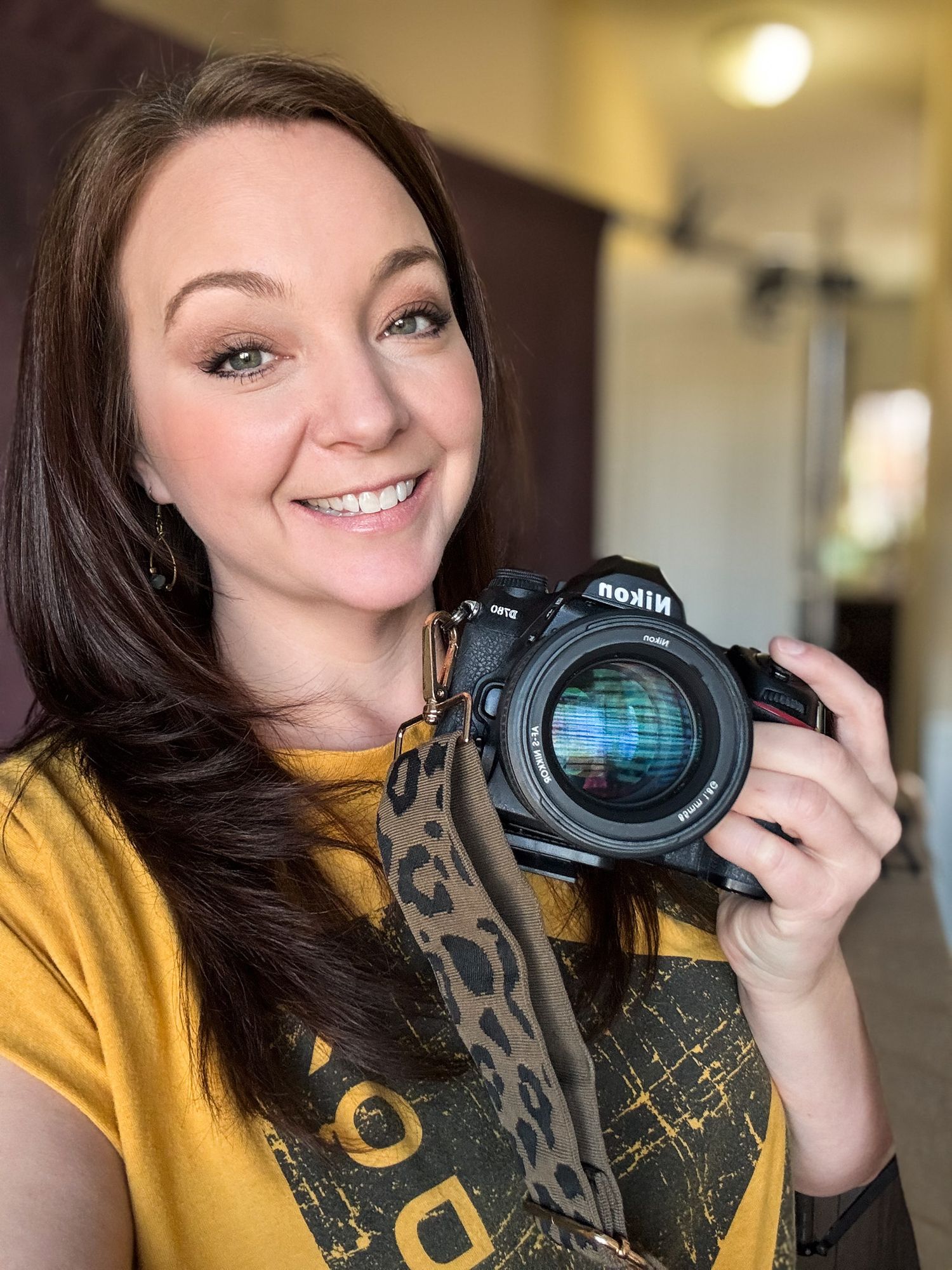 A photographer smiling while holding a Nikon DSLR camera with a lens, wearing a yellow sweater.