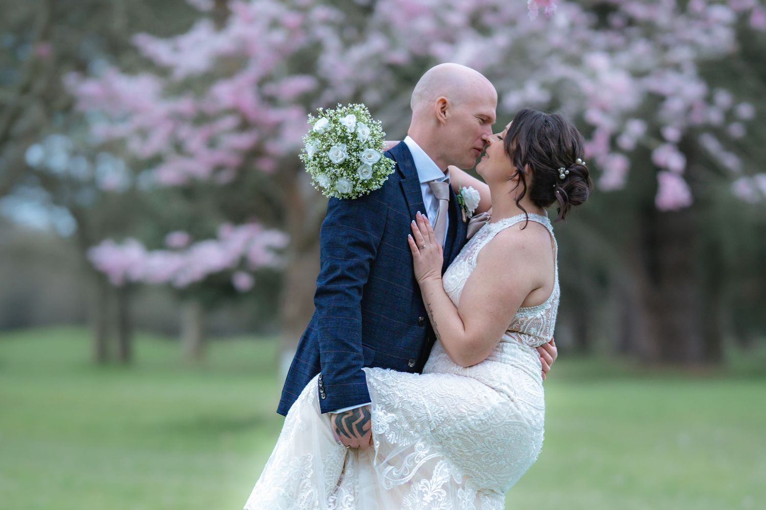 A romantic wedding couple embraces under blooming cherry trees while the bride holds a light blue bouquet.