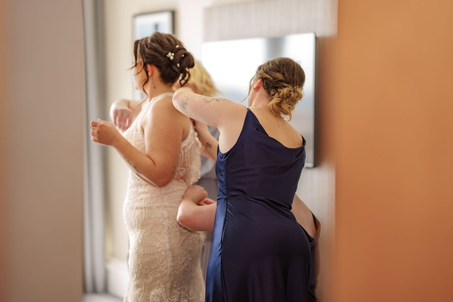 Two people prepare for a wedding ceremony as someone helps secure a white lace dress in a sunlit room.