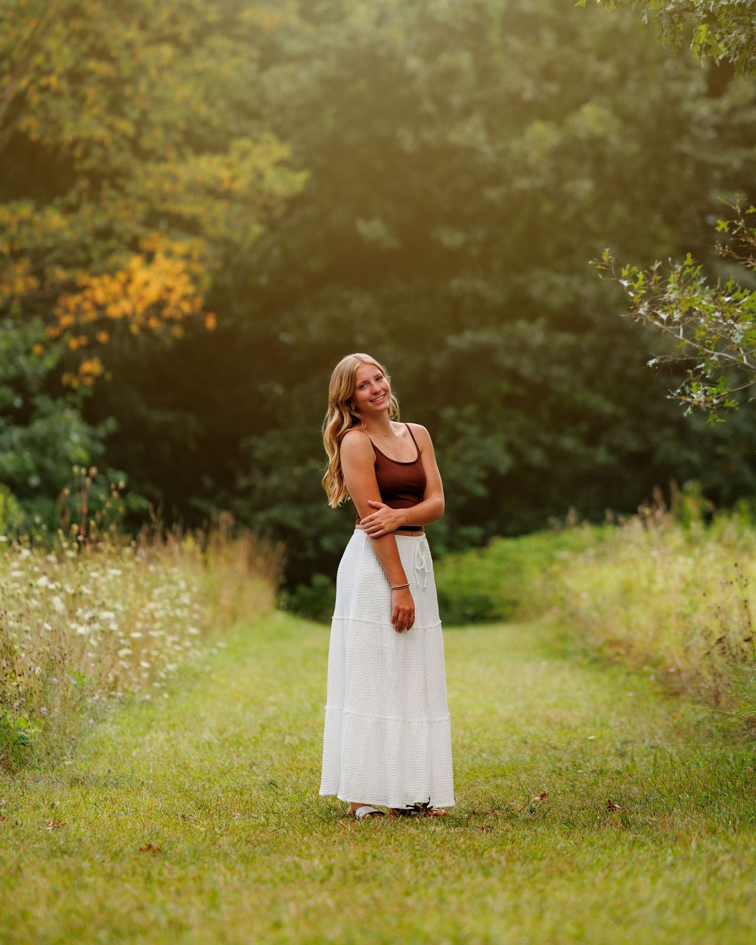 Young woman in white maxi skirt and burgundy top standing on a lush green path surrounded by trees.