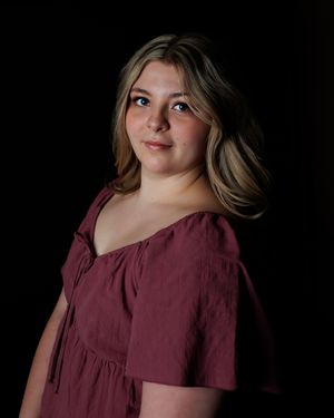 Studio portrait in dramatic lighting against black background showing person in burgundy peasant-style top with flowing hair.