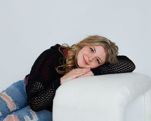 A woman in a black knit sweater relaxes on a white couch wearing distressed jeans.
