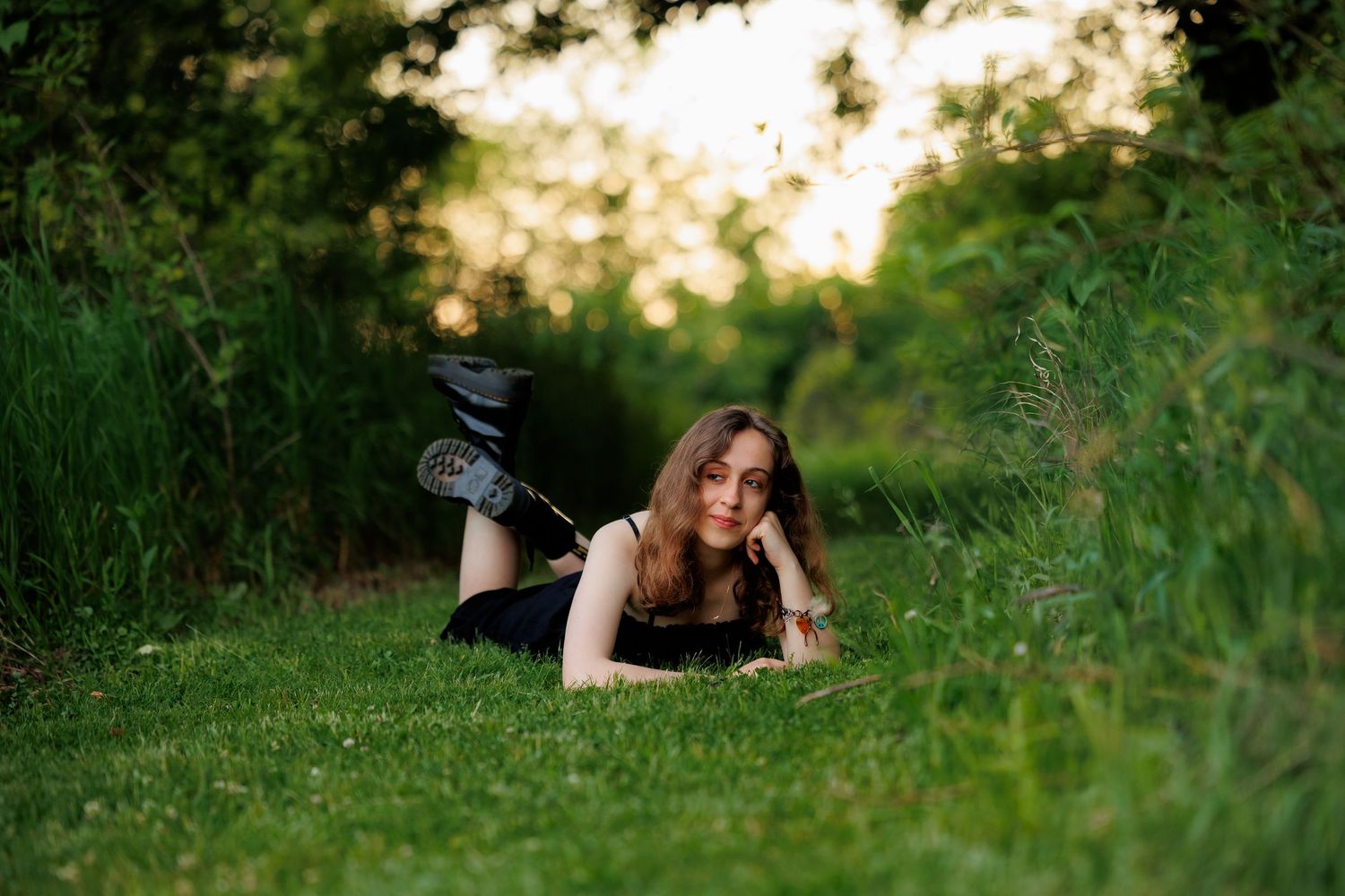 Young woman in black outfit lying on green grass in a sunlit park, resting her chin on her hand.