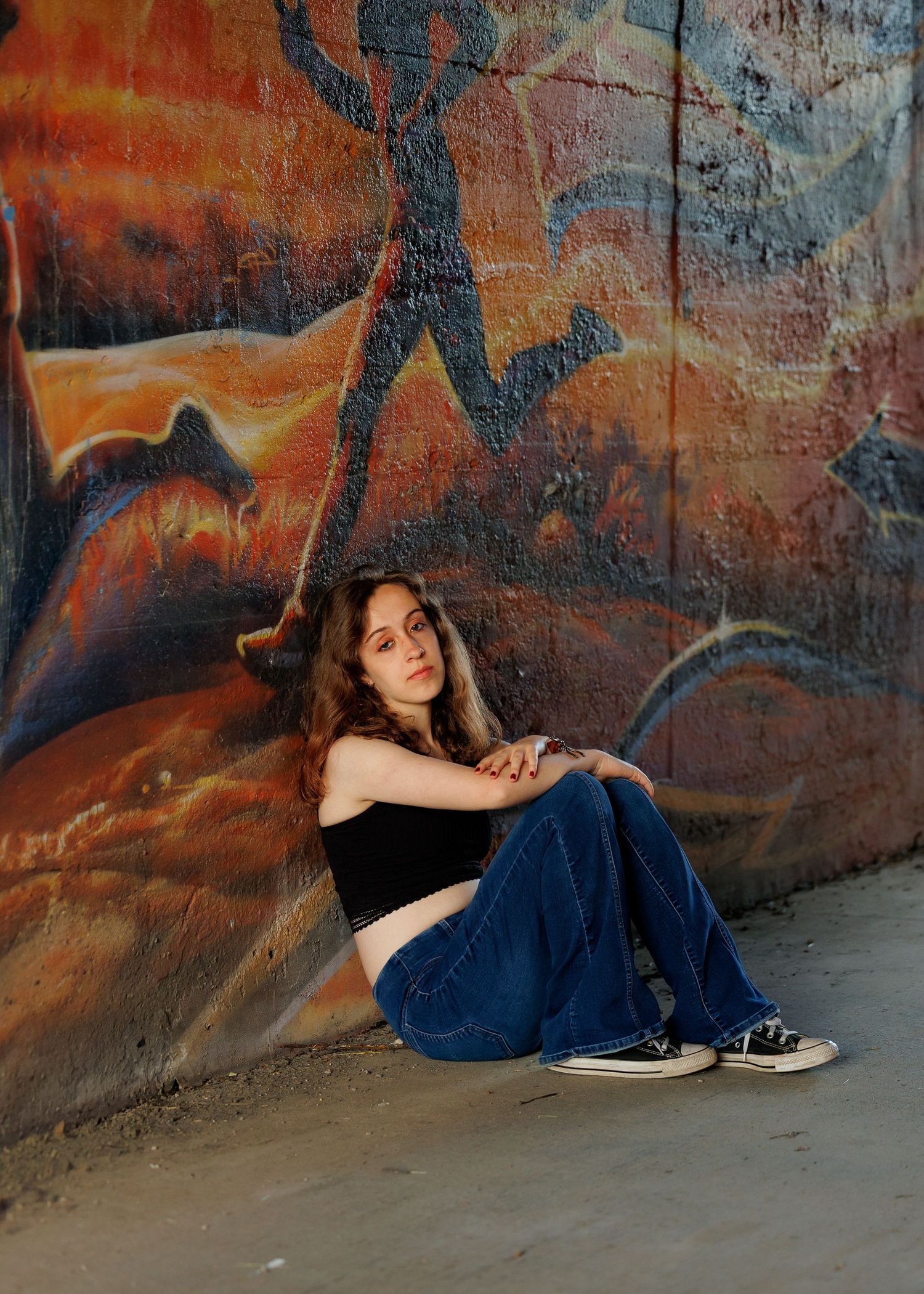Young woman sitting against colorful graffiti wall wearing black crop top, blue jeans, and Converse sneakers.